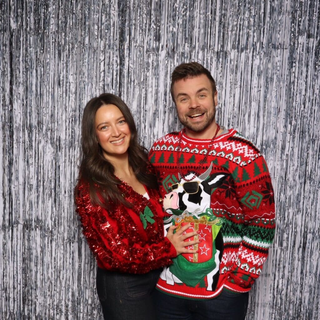 Two guests in ugly Christmas sweaters posing against a silver backdrop at a corporate holiday party photo booth in Kansas City