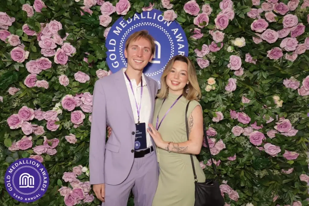 Two guests pose and smile in front of a lush floral wall backdrop featuring mauve roses and greenery at the 53rd Annual Gold Medallion Awards print booth activation in Denver.