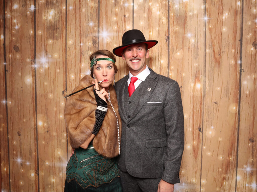 A couple in 1920s Gatsby attire, one in a green beaded dress with fur stole and the other in a grey three-piece suit and black hat, pose against a warm wood backdrop at the St. James Auction, a themed photo experience by Lumen Activations.