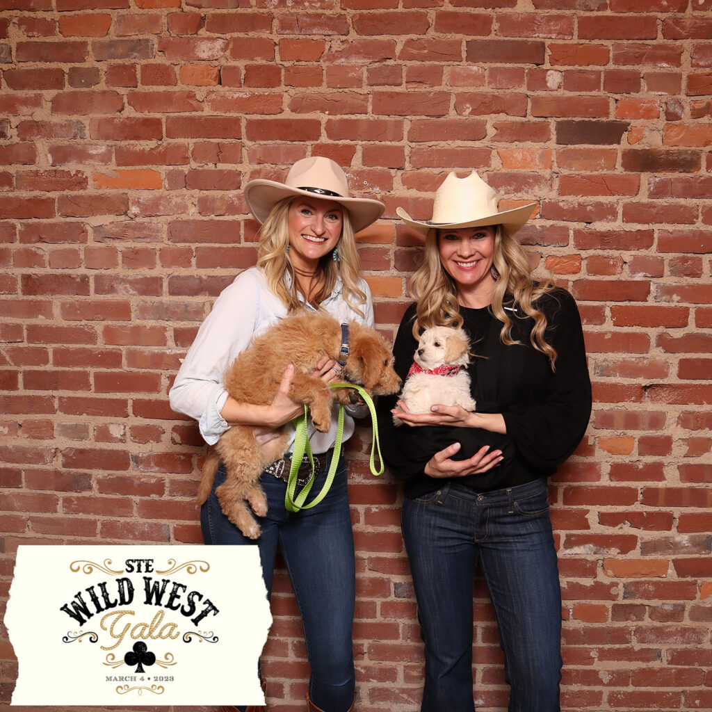 Two guests in cowboy hats and denim hold golden doodle puppies against an exposed brick wall in a square print from the STE Wild West Gala, a themed nonprofit fundraiser photo experience by Lumen Activations.