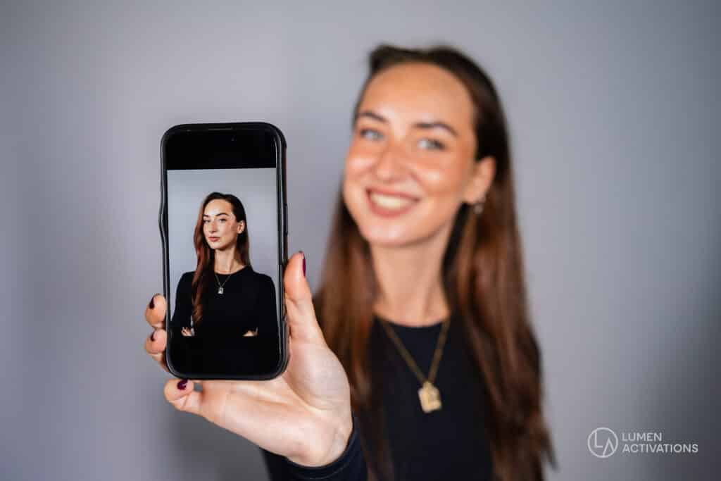 A woman holding a phone showing her professional headshot, captured in a modern studio setup by Lumen Activations.