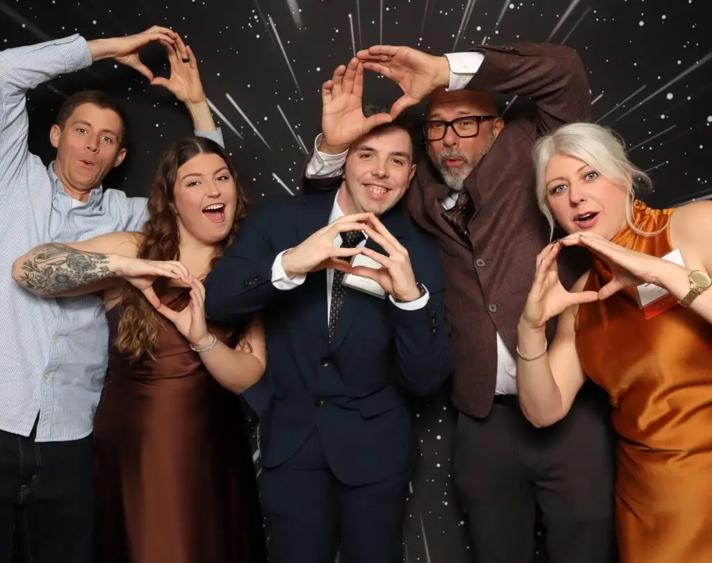 Five coworkers make hand hearts at a corporate celebration photo booth in Kansas City with a starfield backdrop.
