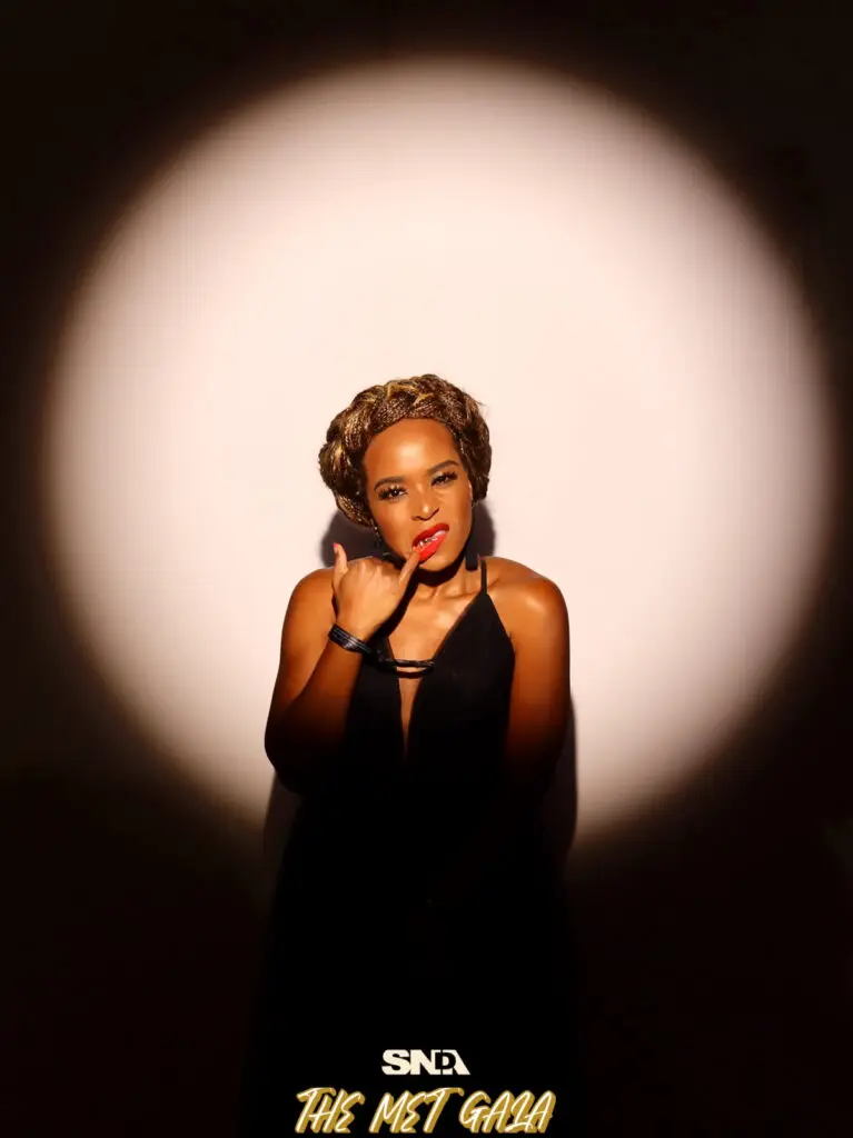 A woman in a black gown with gold braided hair poses under a dramatic spotlight at the SNDA Met Gala "Superfine" photo activation.