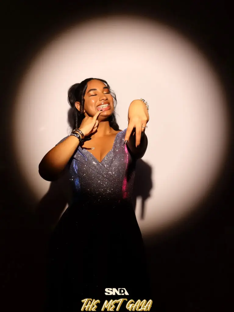 A woman in a sparkling sequin gown strikes a playful pose under a dramatic spotlight at the SNDA Met Gala "Superfine" photo activation.