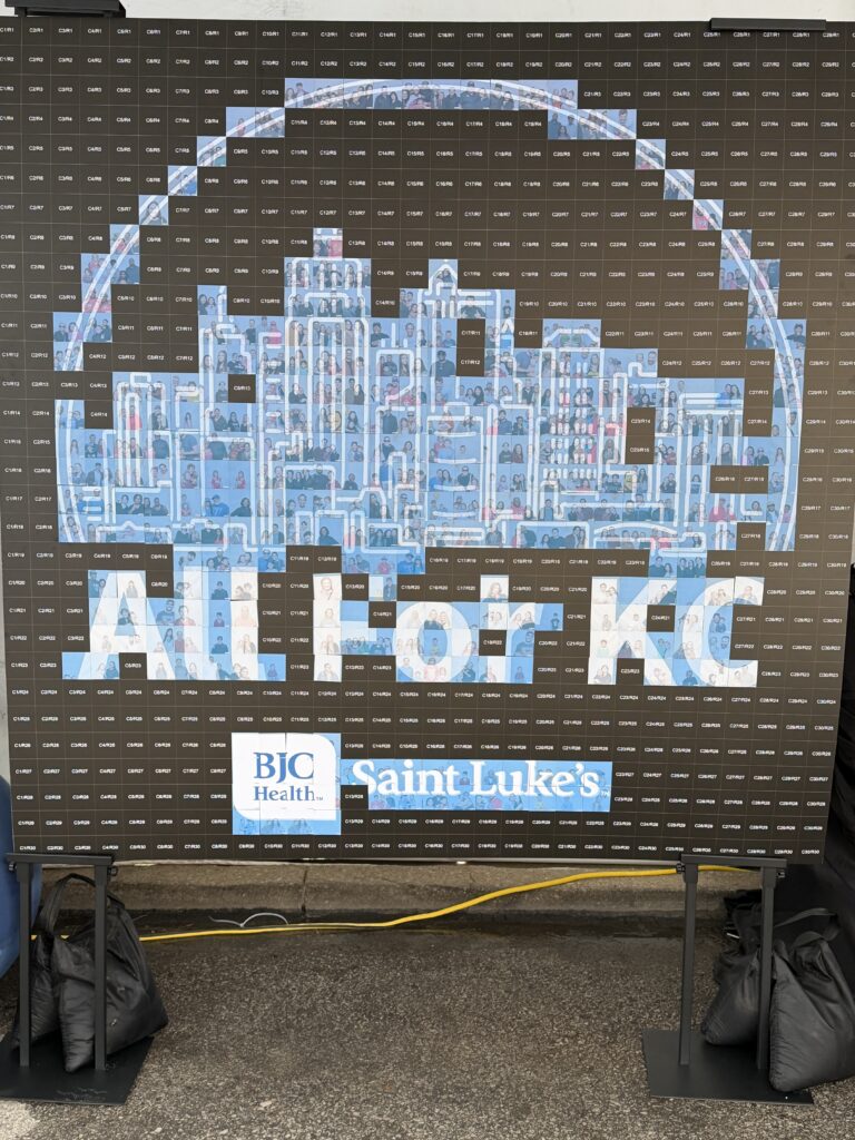 saint lukes photo mosaic wall in progress plaza art fair Saint Luke's All For KC photo mosaic wall partway through the Plaza Art Fair, with guest photo tiles filling the Kansas City skyline and numbered grid squares still waiting to be placed.